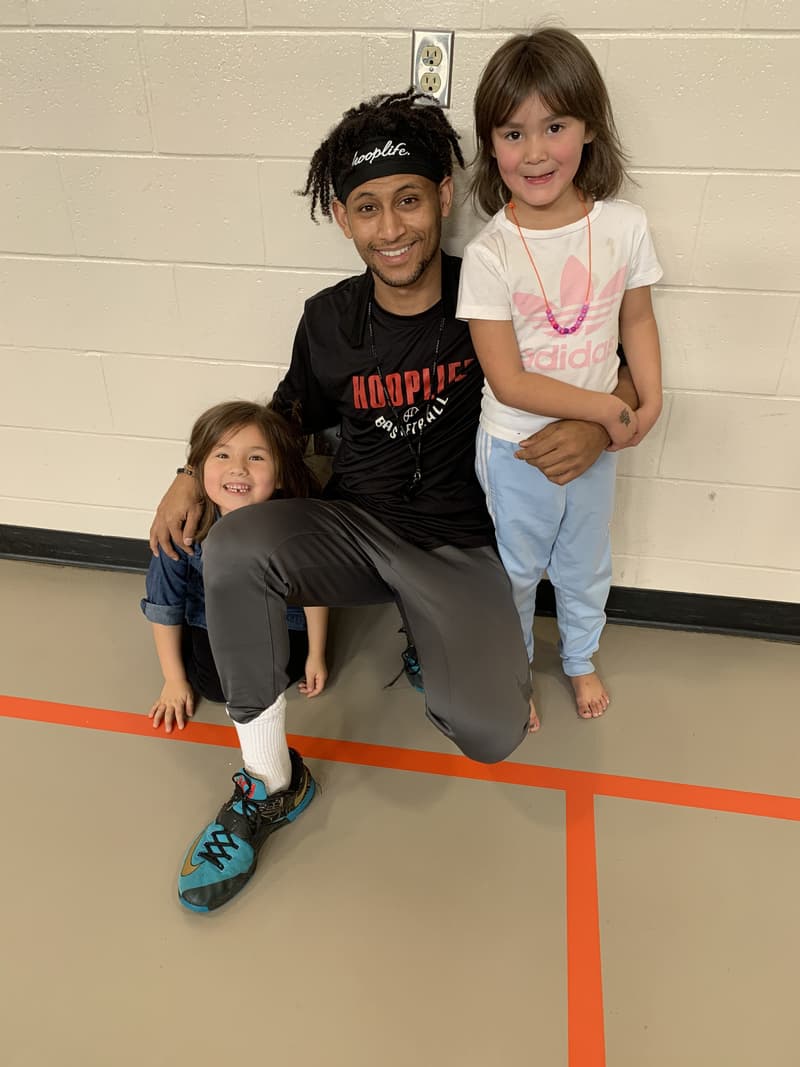 A HoopLife coach kneeling with two smiling young girls at the Whitecap clinic