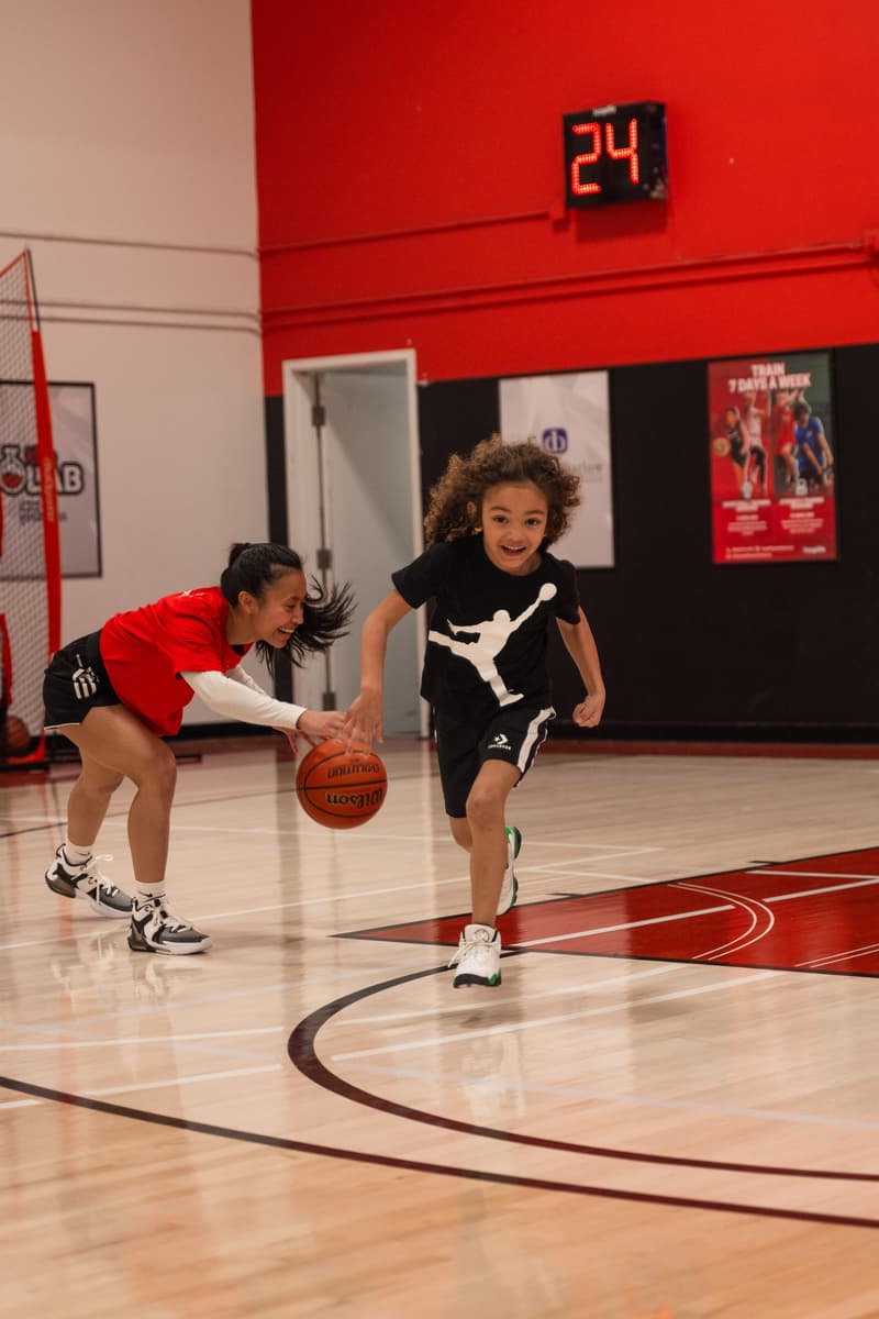 A HoopLife coach dribbling low as a laughing Young Stars kid chases her
